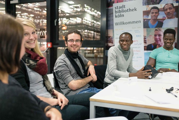 Menschen unterschiedlicher Hautfarbe sitzen im sprachraum in der Stadtbibliothek Köln um einen Tisch herum und reden miteinander.