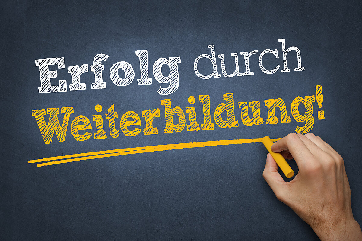 Auf einer Tafel steht "Erfolg durch Weiterbildung". Eine Hand mit Kreide unterstreicht das Wort "Weiterbildung".